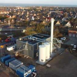 The picture shows the new EVS CHP plant in Westerland. In front of the building, the two 100 m³ buffer storage tanks and the 28 m high chimney can be seen. Lying on the roof, the secondary silencers of the two CHP units can be seen.